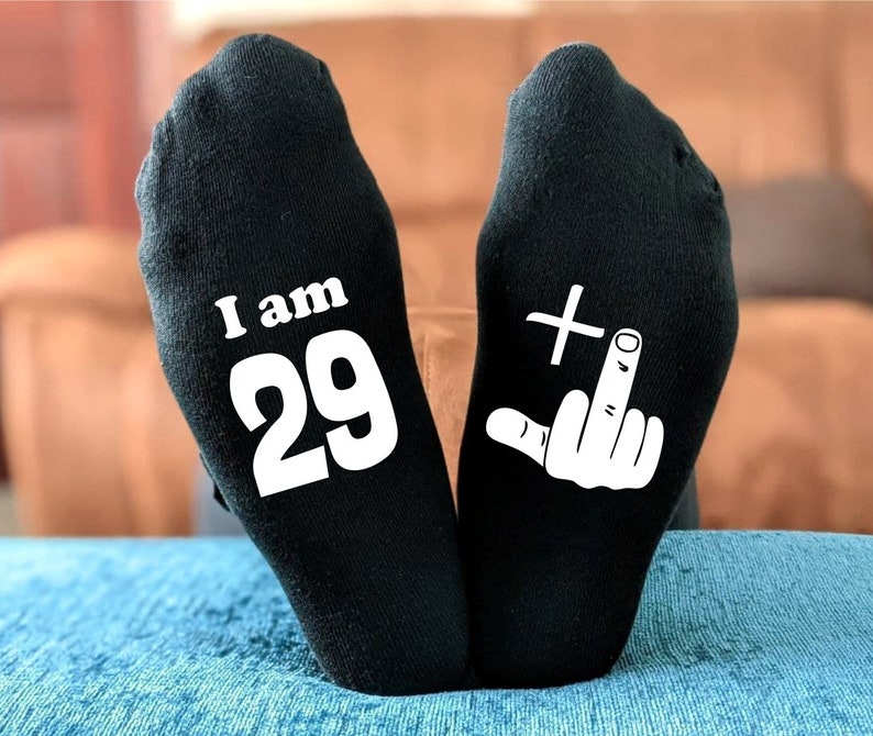 May include: A pair of black socks with white text. One sock says "I am 29" and the other sock has a white middle finger with a plus sign above it.