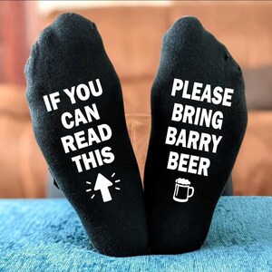 Peut inclure: Paire de chaussettes noires avec du texte blanc. Une chaussette dit "IF YOU CAN READ THIS" avec une flèche, l'autre dit "PLEASE BRING BARRY BEER" avec un dessin de chope de bière. Les chaussettes sont posées sur une surface bleue.