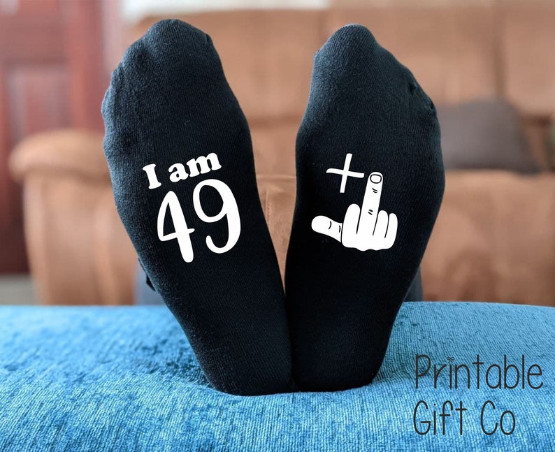 May include: A pair of black socks with white text. One sock says "I am 49" and the other sock has a white plus sign and a middle finger.