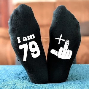 May include: A pair of black socks with white text. One sock says "I am 79" and the other sock has a white middle finger with a plus sign above it.