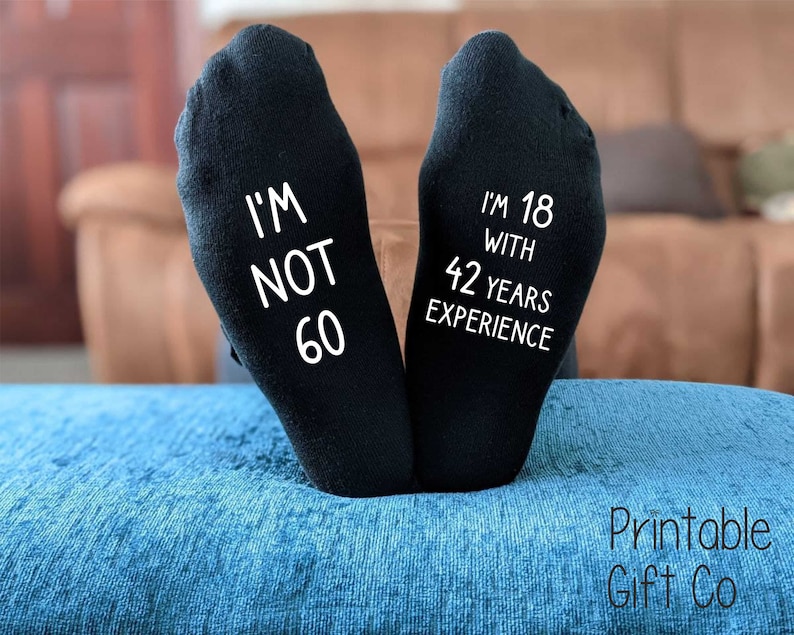 May include: A pair of black socks with white text. One sock reads "I'm not 60" and the other reads "I'm 18 with 42 years experience".