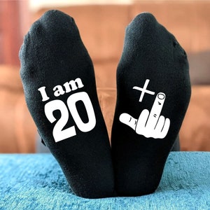 May include: A pair of black socks with white text. One sock says "I am 20" and the other sock has a white middle finger with a plus sign above it.