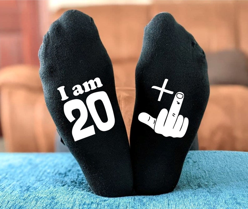 May include: A pair of black socks with white text. One sock says "I am 20" and the other sock has a white middle finger with a plus sign above it.