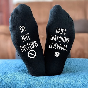 May include: A pair of black socks with white text. One sock says "Do Not Disturb" with a no entry sign. The other sock says "Dad's Watching Liverpool" with a football.