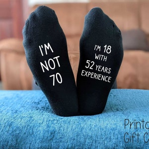 May include: A pair of black socks with white text. One sock says "I'm not 70" and the other sock says "I'm 18 with 52 years experience".