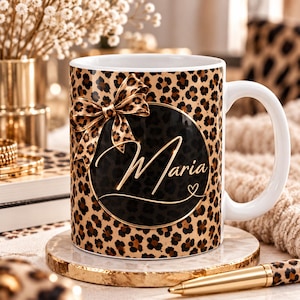 Può includere: Una tazza in ceramica bianca con motivo leopardato marrone e nero. Un emblema circolare nero centrale mostra il nome Maria in corsivo dorato, accentuato da un fiocco leopardato con bordo dorato sopra il cerchio.
