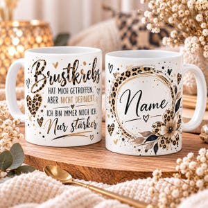 May include: Two white ceramic mugs featuring gold and black leopard print accents. One mug displays the German text "Brustkrebs hat mich getroffen, aber nicht definiert. Ich bin immer noch ich. Nur stärker." The other mug features a leopard print circle frame with the word "Name" and a floral design.