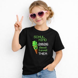 Könnte beinhalten: Schwarzes T-Shirt mit einem grünen, pixeligen Design, das eine Eistüte mit einem Charakter im Minecraft-Stil und dem Text "SCHUL KIND 2026 Next Level THEA" zeigt. Das Mädchen trägt eine rosa herzförmige Sonnenbrille und zeigt das Peace-Zeichen.