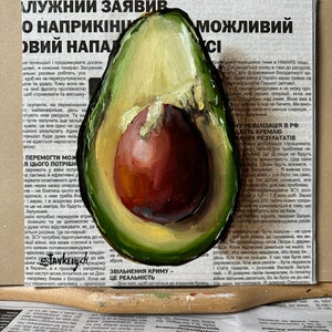 May include: A painting of a halved avocado with a brown pit and a green outer layer. The avocado is painted on top of a newspaper article with text in Ukrainian.