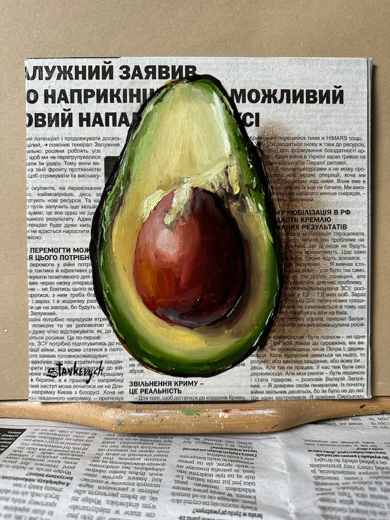 May include: A painting of a halved avocado with a brown pit and a green outer layer. The avocado is painted on top of a newspaper article with text in Ukrainian.