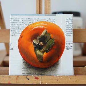 Puede incluir: Pintura al &oacute;leo de un caqui naranja brillante con hojas verdes sobre lienzo. La obra de arte est&aacute; sobre un caballete de madera. El fondo presenta texto de peri&oacute;dico.