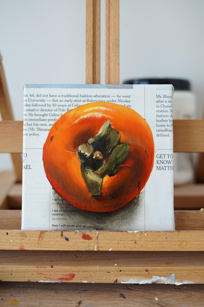 Puede incluir: Pintura al &oacute;leo de un caqui naranja brillante con hojas verdes sobre lienzo. La obra de arte est&aacute; sobre un caballete de madera. El fondo presenta texto de peri&oacute;dico.