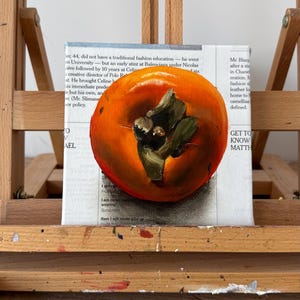 Puede incluir: Pintura al &oacute;leo de un caqui maduro con tallo y hojas verdes, sobre un fondo de texto de peri&oacute;dico. La fruta es de color naranja brillante, con pinceladas detalladas.
