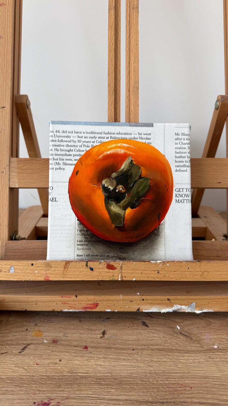 Puede incluir: Pintura al &oacute;leo de un caqui maduro con tallo y hojas verdes, sobre un fondo de texto de peri&oacute;dico. La fruta es de color naranja brillante, con pinceladas detalladas.