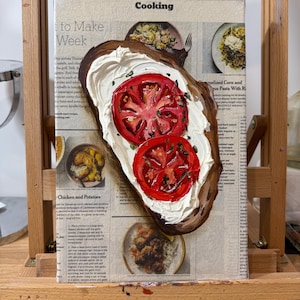 May include: A painting of a slice of toasted bread topped with cream cheese and two red tomato slices. The painting is on a canvas with a newspaper background that includes the text "Cooking" and "Chicken and Potatoes".