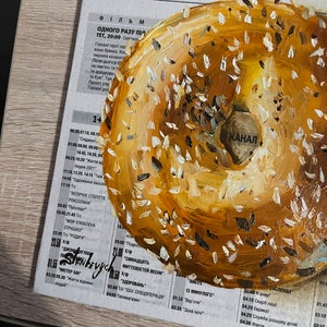 May include: A painting of a bagel with sesame seeds. The bagel is painted in shades of brown and yellow. The background is a newspaper with text in Ukrainian.