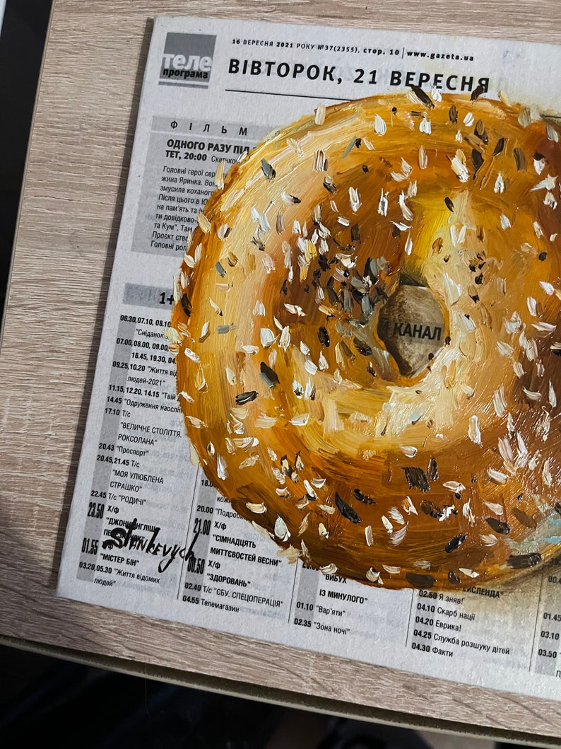 May include: A painting of a bagel with sesame seeds. The bagel is painted in shades of brown and yellow. The background is a newspaper with text in Ukrainian.