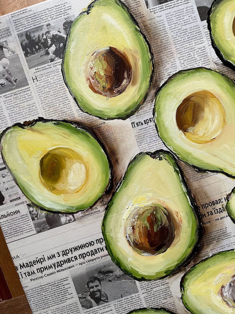 May include: A painting of several avocado halves on a newspaper background. The avocados are painted in a realistic style with green skin and yellow flesh. The newspaper is blurred and unreadable.