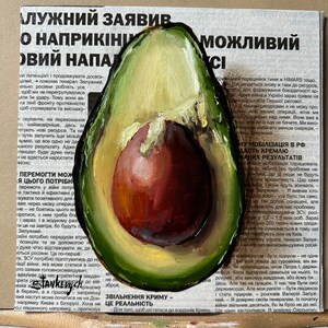 May include: A close-up of a painting of a halved avocado with a red pit. The avocado is painted in a realistic style with green and yellow hues. The pit is painted in a deep red color. The painting is on a canvas with a newspaper background. The newspaper text is partially visible and reads "ZAVIV O NAPRIKINI OVIY NAPAD" and "ZVILNENNIA KRIMU - CE REALNIST".