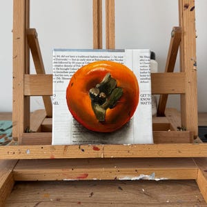 Puede incluir: Pintura de naturaleza muerta de un caqui sobre un lienzo, apoyada en un caballete de madera. La fruta es de color naranja vibrante con hojas verdes. El lienzo est&aacute; colocado encima de un peri&oacute;dico. El caballete es de madera marr&oacute;n claro.