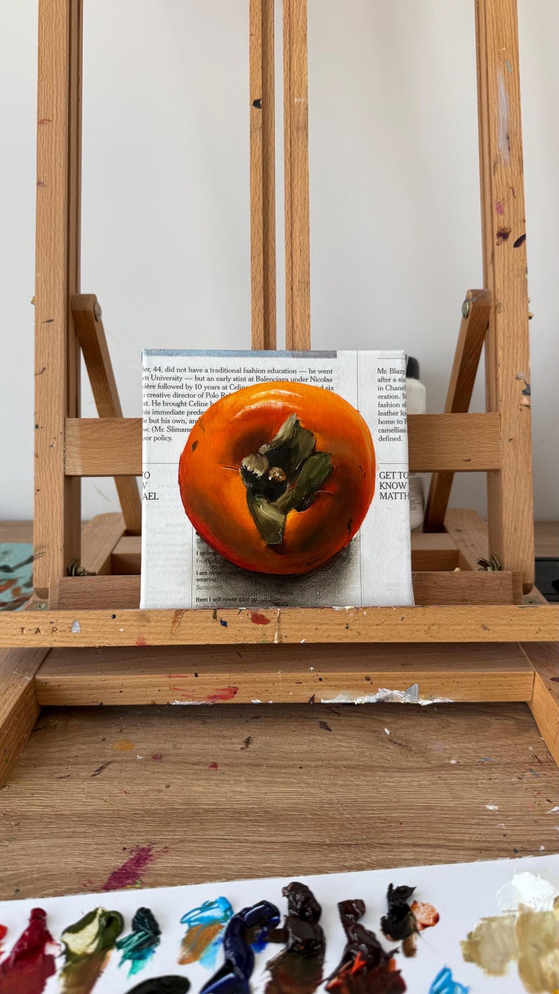 Puede incluir: Pintura de naturaleza muerta de un caqui sobre un lienzo, apoyada en un caballete de madera. La fruta es de color naranja vibrante con hojas verdes. El lienzo est&aacute; colocado encima de un peri&oacute;dico. El caballete es de madera marr&oacute;n claro.