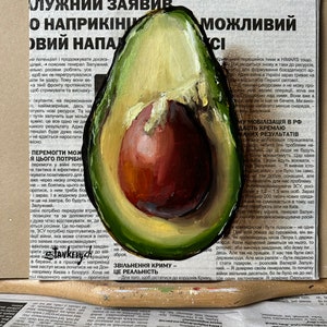 May include: A painting of a halved avocado with a green outer skin and a brown pit. The avocado is painted on top of a newspaper article with the text "ZVILNENNIA KRYMU - CE REALNIST".