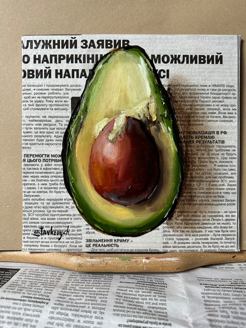 May include: A painting of a halved avocado with a green outer skin and a brown pit. The avocado is painted on top of a newspaper article with the text "ZVILNENNIA KRYMU - CE REALNIST".