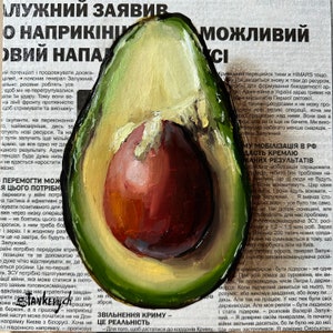 May include: A close-up of a halved avocado with a large seed. The avocado is painted in a realistic style with green, brown, and yellow hues. The seed is painted in a reddish-brown color. The avocado is sitting on top of a newspaper article with text in a foreign language.