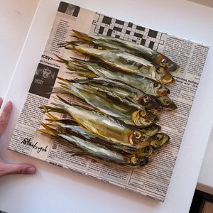 May include: A painting of a still life featuring a group of silver fish arranged on top of a newspaper. The fish are painted in a realistic style with details of their scales and fins. The newspaper is partially visible in the background, with some text visible.