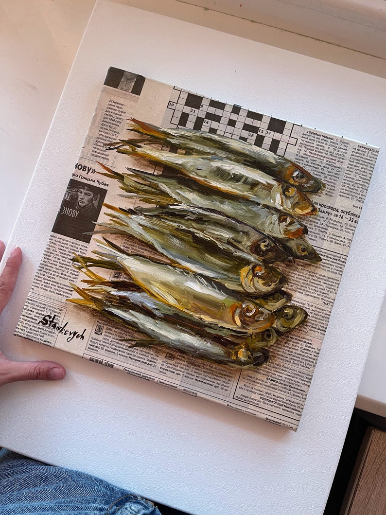 May include: A painting of a still life featuring a group of silver fish arranged on top of a newspaper. The fish are painted in a realistic style with details of their scales and fins. The newspaper is partially visible in the background, with some text visible.