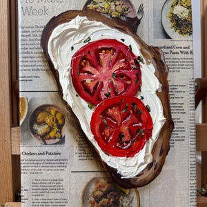May include: A painting of a slice of toasted bread topped with white cream cheese and two red tomato slices. The painting is on a canvas with a newspaper clipping visible in the background. The newspaper clipping is about cooking and features a recipe for chicken and potatoes.