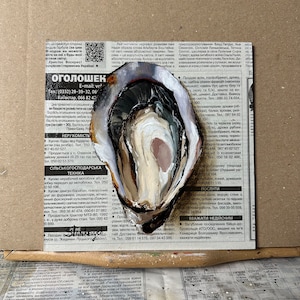 May include: A close-up of a white oyster shell with a dark brown interior. The oyster shell is painted with white, brown, and pink paint. The oyster shell is sitting on a newspaper with text in Ukrainian.