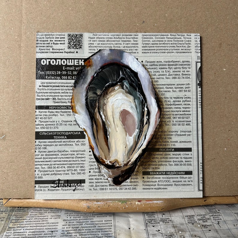 May include: A close-up of a white oyster shell with a dark brown interior. The oyster shell is painted with white, brown, and pink paint. The oyster shell is sitting on a newspaper with text in Ukrainian.