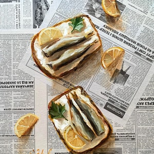 May include: Two open-faced sandwiches with sardines, lemon slices, and parsley on a newspaper background.