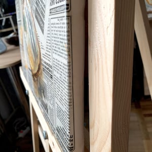 May include: A wooden easel with a canvas stretched over it. The canvas is covered in newspaper clippings with text in a foreign language. The text on the canvas reads "ВИПАДОКИ АРОДИЛАС WHA in нар буг ト бута HE У ас народилася п ма: знати, що хочек арабул ЖИТЕВА ІСТОРІЯ ЕРЕЗ АКУ ПОДРУЖ".