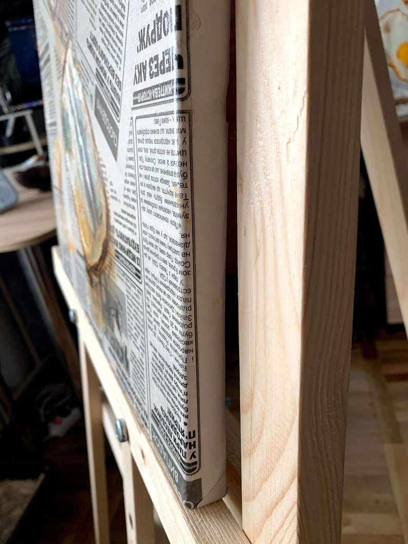 May include: A wooden easel with a canvas stretched over it. The canvas is covered in newspaper clippings with text in a foreign language. The text on the canvas reads "ВИПАДОКИ АРОДИЛАС WHA in нар буг ト бута HE У ас народилася п ма: знати, що хочек арабул ЖИТЕВА ІСТОРІЯ ЕРЕЗ АКУ ПОДРУЖ".