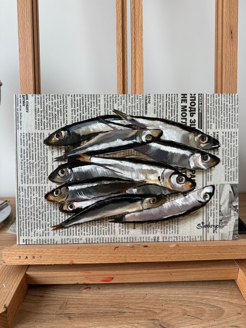 May include: A painting of a group of silver fish on a newspaper background. The fish are arranged in a pile, with their tails overlapping. The newspaper is printed in black and white, and the text is visible. The painting is done in a realistic style, with the fish appearing to be three-dimensional.