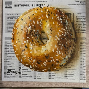 May include: A painting of a round, sesame seed bagel with a brown and white glaze. The bagel is painted on a newspaper with a Ukrainian television program schedule. The text on the newspaper reads "ВІВТОРОК, 21 ВЕРЕСНЯ" which translates to "Tuesday, September 21st".