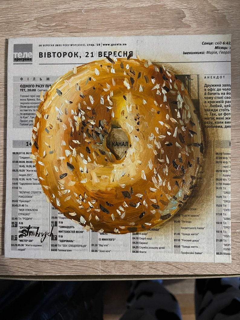 May include: A painting of a round, sesame seed bagel with a brown and white glaze. The bagel is painted on a newspaper with a Ukrainian television program schedule. The text on the newspaper reads "ВІВТОРОК, 21 ВЕРЕСНЯ" which translates to "Tuesday, September 21st".