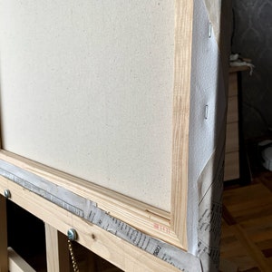 May include: A close-up of the corner of a wooden canvas frame. The canvas is stretched tightly over the frame and is secured with staples. The frame is made of natural wood and has a light brown finish. The canvas is white and has a fine texture.
