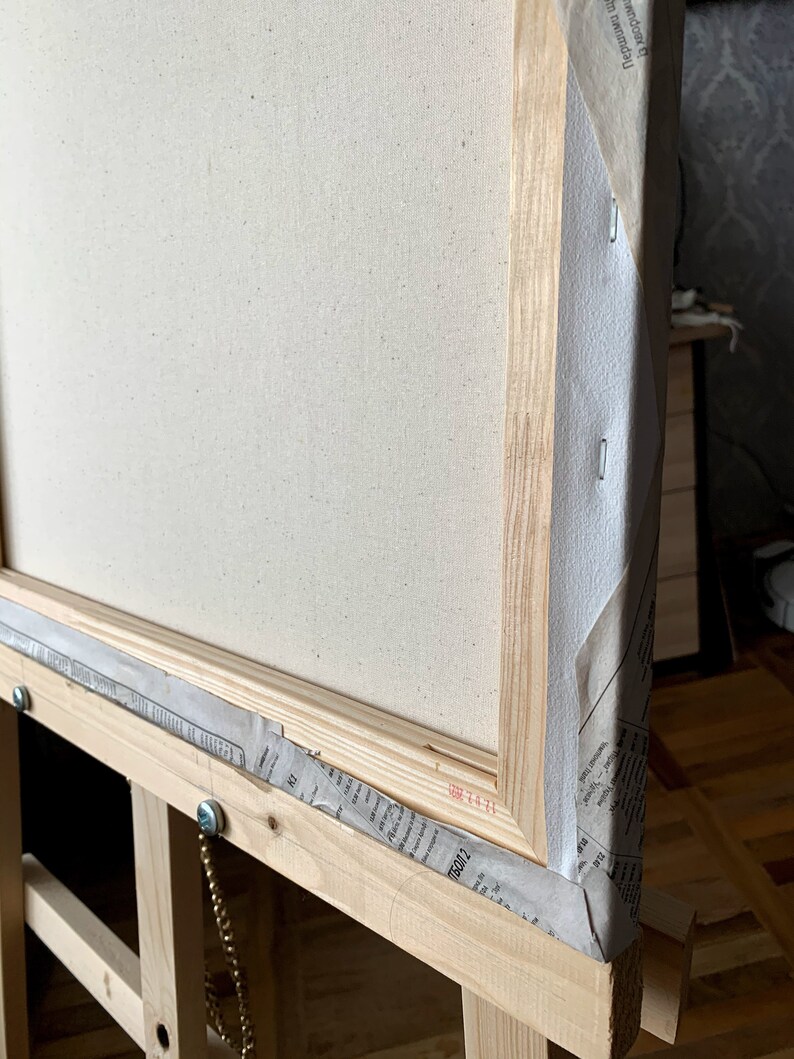 May include: A close-up of the corner of a wooden canvas frame. The canvas is stretched tightly over the frame and is secured with staples. The frame is made of natural wood and has a light brown finish. The canvas is white and has a fine texture.