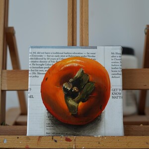 Puede incluir: Pintura al &oacute;leo de un caqui maduro, en tonos naranja y verde, sobre un fondo de texto de peri&oacute;dico. La obra de arte se exhibe en un caballete de madera, mostrando la textura y la forma de la fruta.