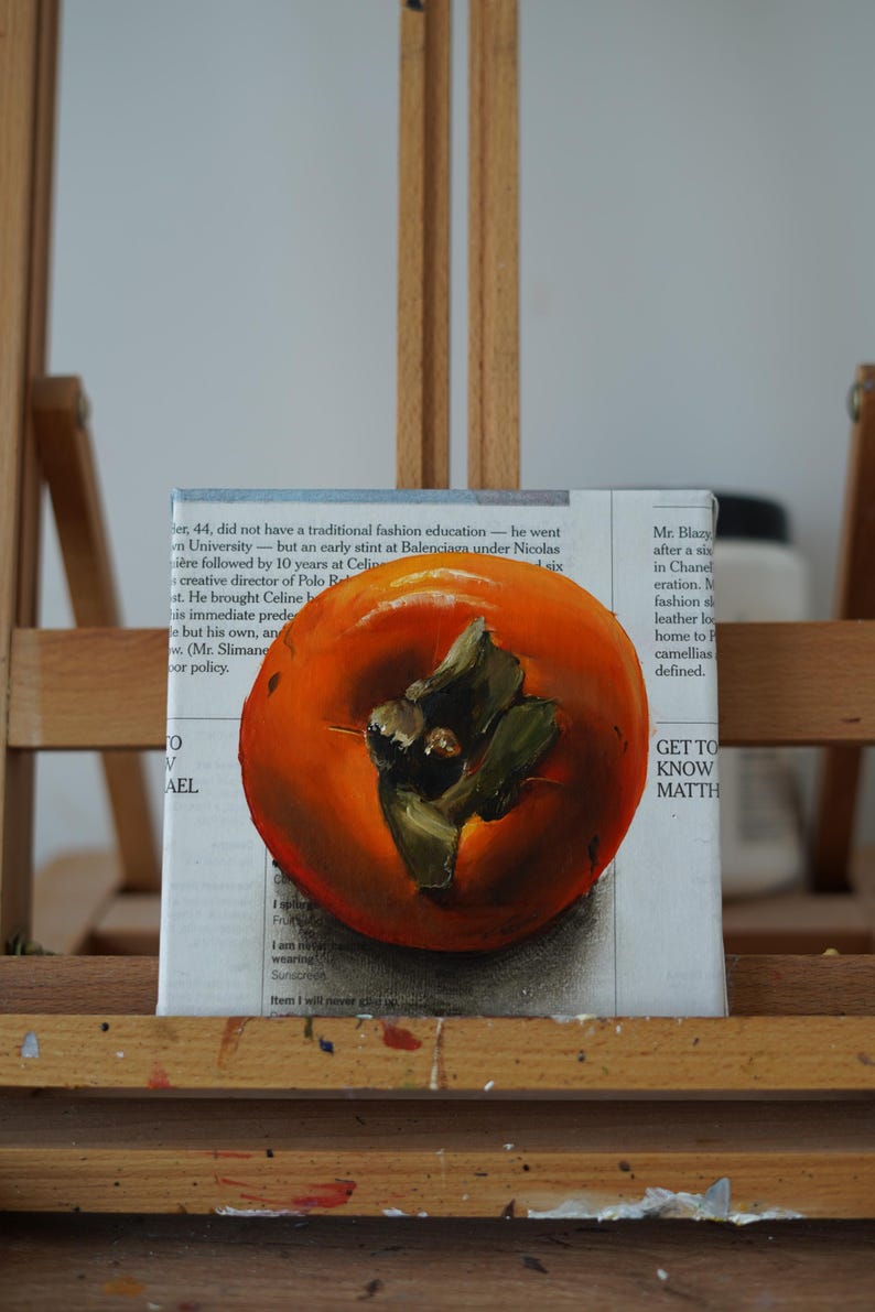 Puede incluir: Pintura al &oacute;leo de un caqui maduro, en tonos naranja y verde, sobre un fondo de texto de peri&oacute;dico. La obra de arte se exhibe en un caballete de madera, mostrando la textura y la forma de la fruta.
