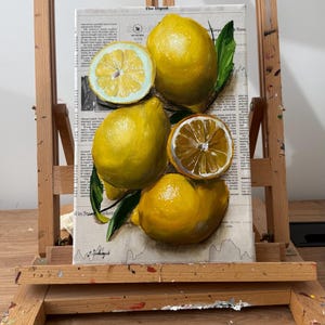 May include: A rectangular canvas painting featuring four bright yellow lemons, two of which are sliced in half, with green leaves. The lemons are painted over a background of printed newspaper text titled The Digest, with a signature at the bottom.