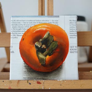 Puede incluir: Pintura al &oacute;leo de un caqui maduro sobre lienzo, expuesta en un caballete de madera. La fruta es de color naranja vibrante con hojas verdes y est&aacute; pintada sobre un lienzo con texto de peri&oacute;dico visible en el fondo.