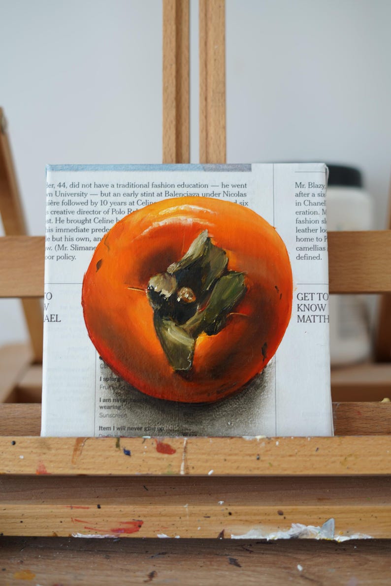 Puede incluir: Pintura al &oacute;leo de un caqui maduro sobre lienzo, expuesta en un caballete de madera. La fruta es de color naranja vibrante con hojas verdes y est&aacute; pintada sobre un lienzo con texto de peri&oacute;dico visible en el fondo.