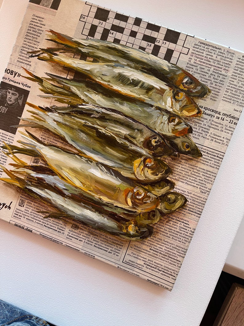 May include: A painting of a pile of dried fish on a newspaper background. The fish are arranged in a vertical stack, with the tails at the top and the heads at the bottom. The fish are a light brown color with a slightly iridescent sheen. The newspaper is a light brown color with black text.