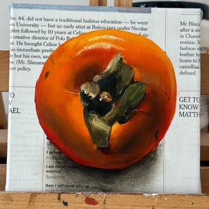Puede incluir: Una pintura al &oacute;leo de un caqui maduro, que muestra tonos naranjas vibrantes con hojas verdes. La obra de arte est&aacute; ambientada sobre un fondo de texto de peri&oacute;dico, montada sobre un caballete de madera, capturando un tema de naturaleza muerta.
