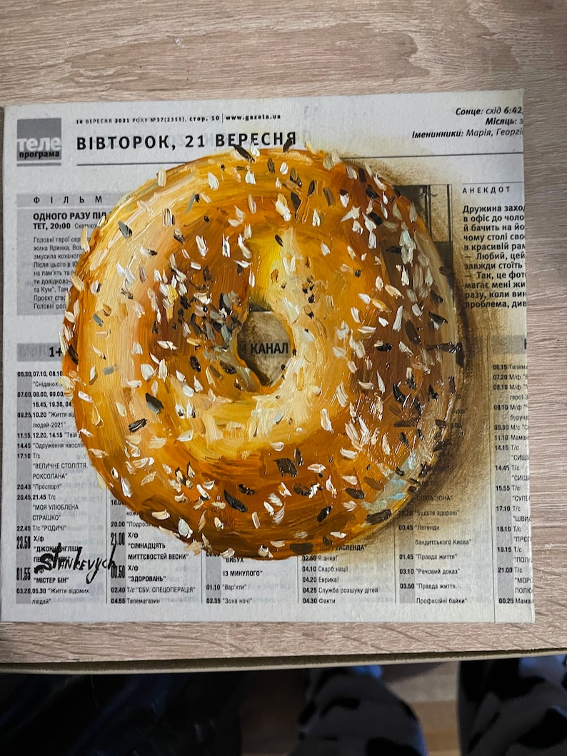 May include: A painting of a bagel with sesame seeds on a newspaper background. The newspaper headline reads "ВІВТОРОК, 21 ВЕРЕСНЯ".