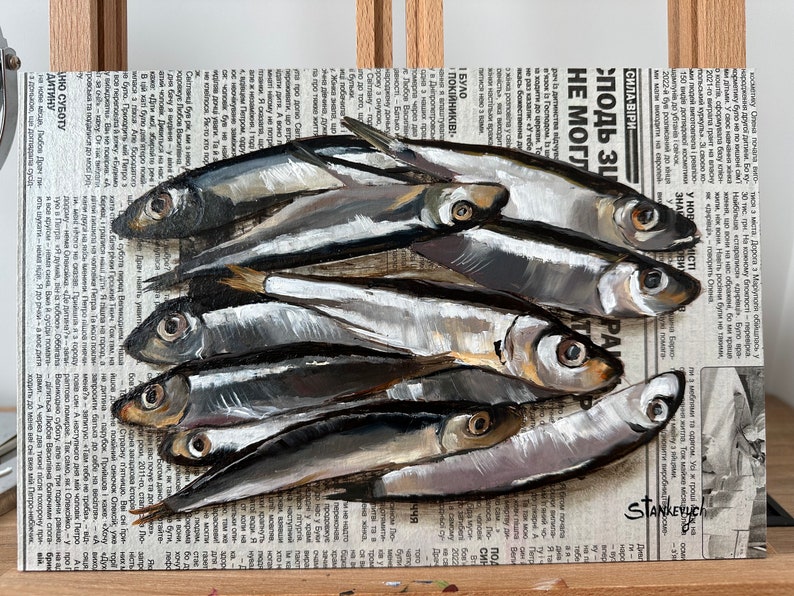 May include: A painting of a group of silver fish on a newspaper background. The fish are arranged in a pile, with their tails pointing towards the top of the image. The newspaper is printed in Russian and has the words "СИЛА-ВІРИ" and "подь Зи" visible. The painting is signed "STANKEYUCH" in the lower right corner.